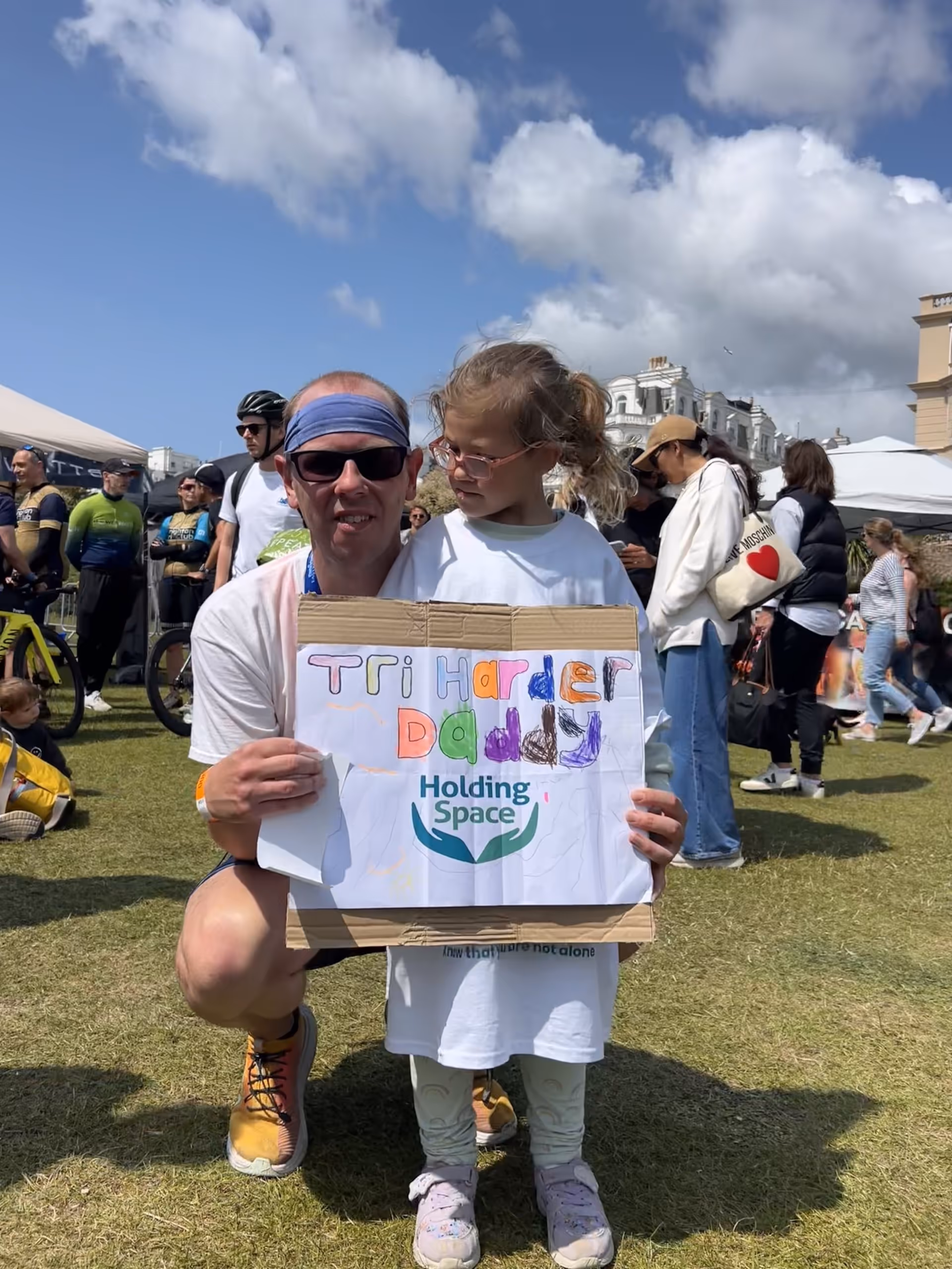Man and child hold a colorful "Tri Harder Daddy" sign at an outdoor event.