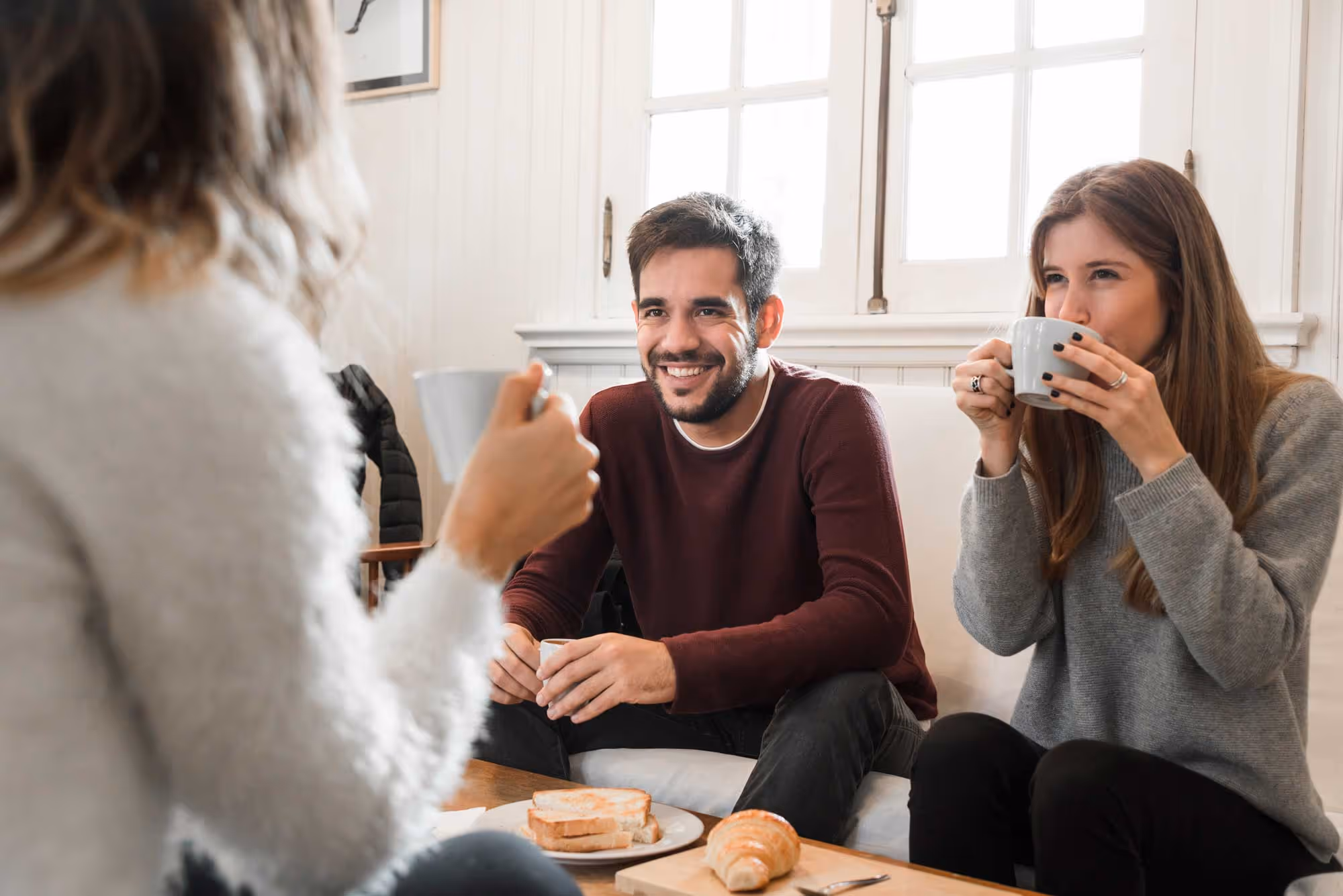 Three people sitting on a couch, smiling and drinking hot beverages, with pastries on a table.