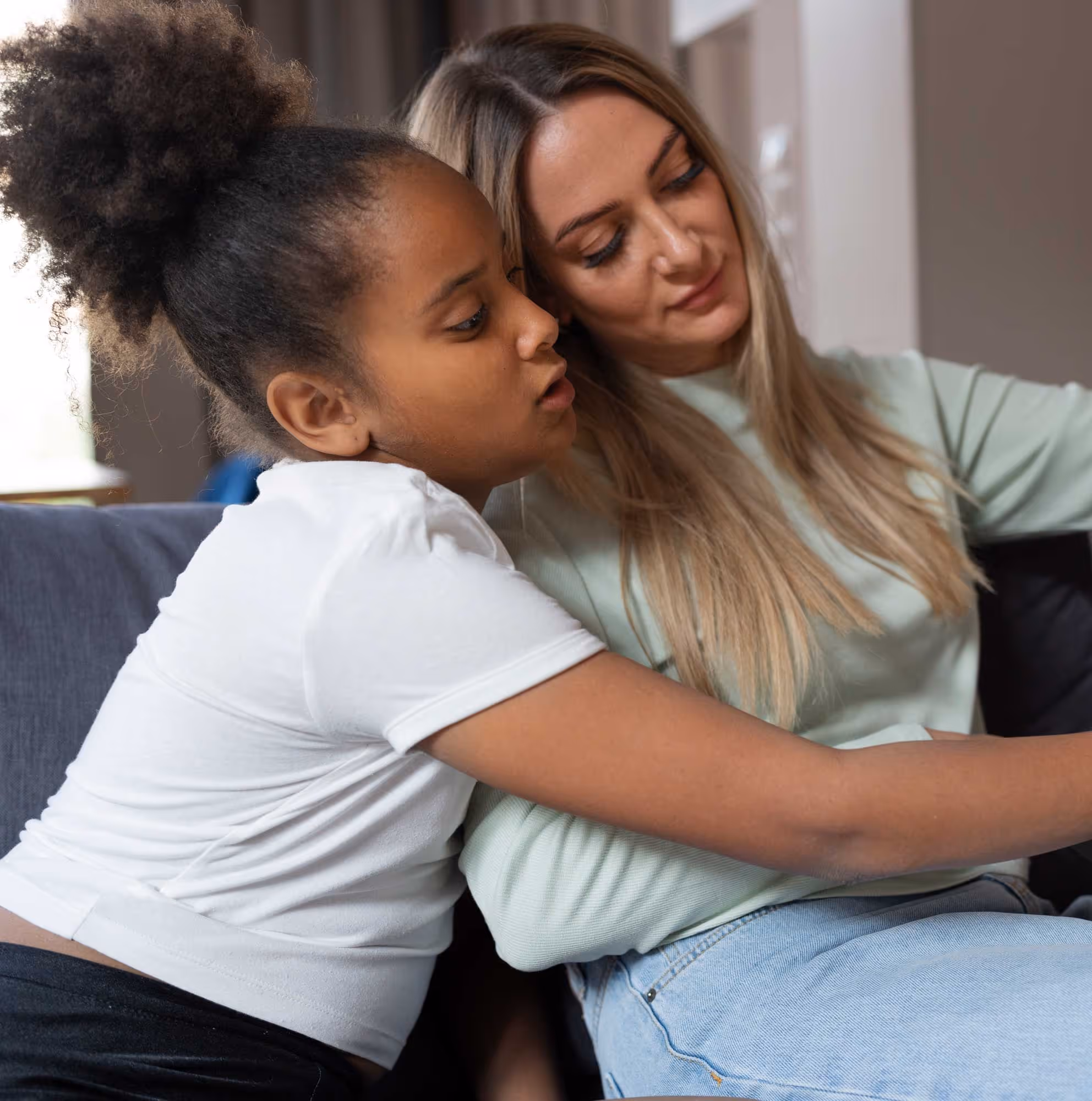 A child with curly hair hugs a woman with long hair while sitting on a couch.