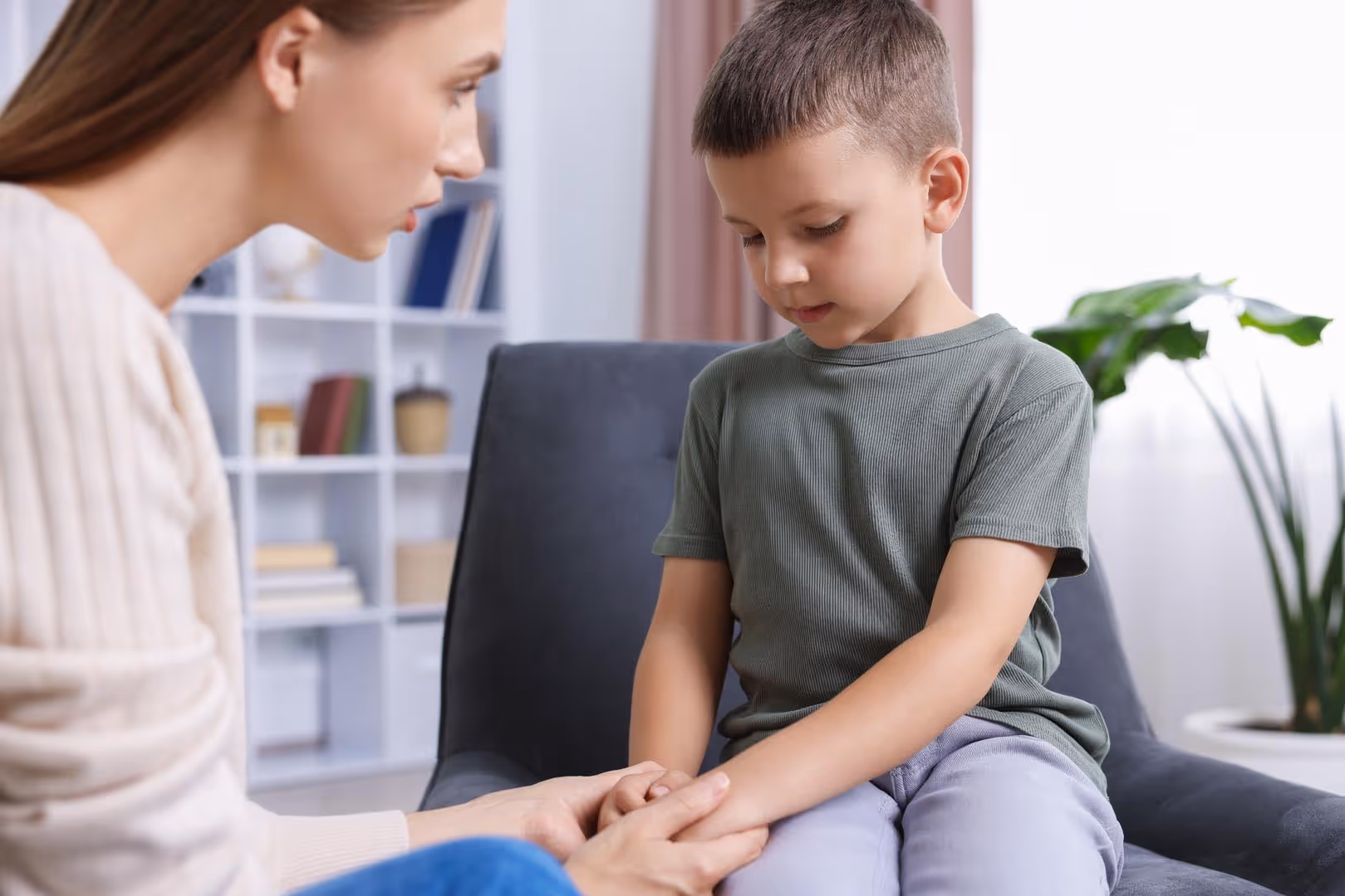 A woman gently holds a boy's hand as they sit together, both looking thoughtful.