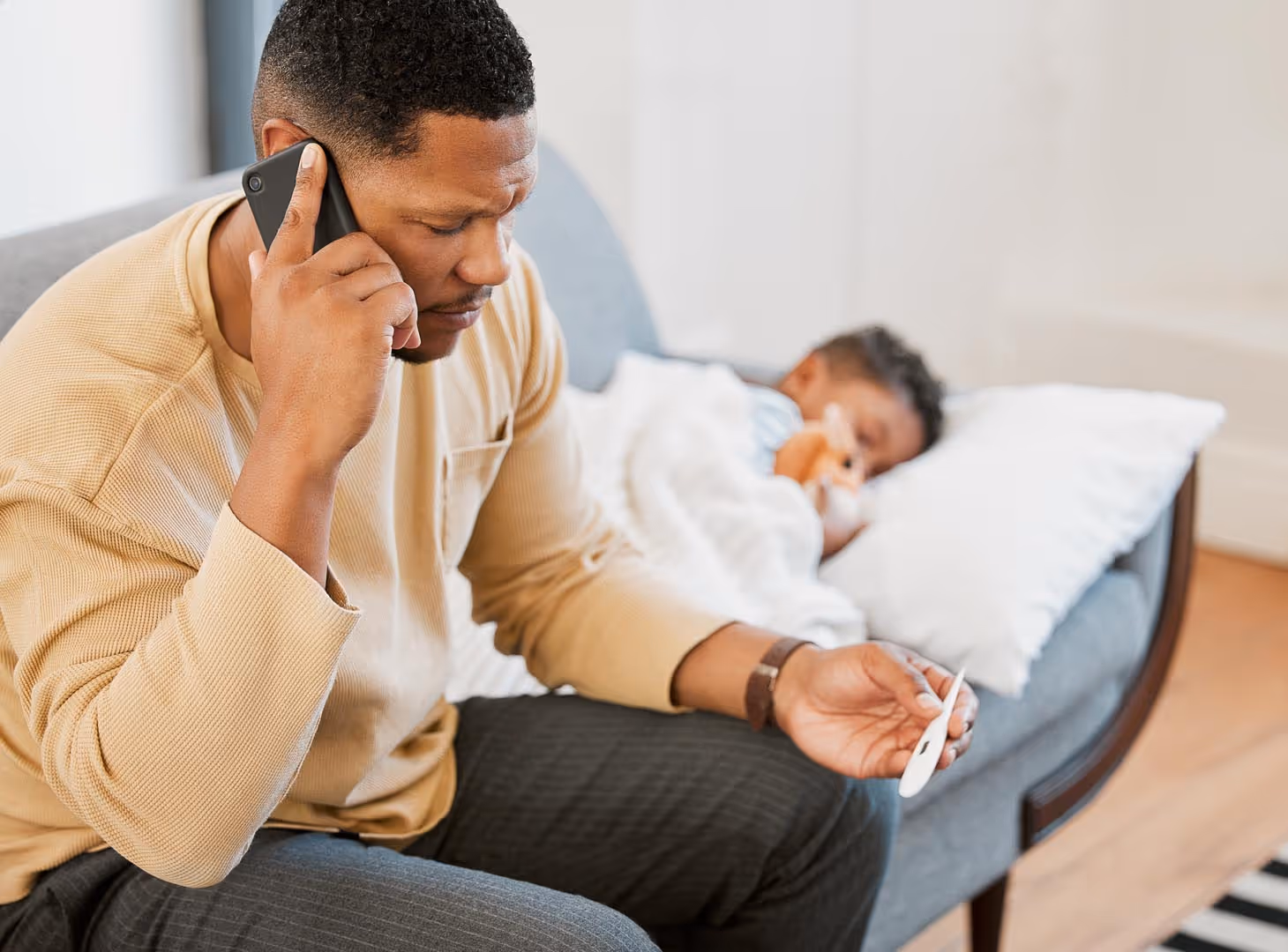 Man on phone checks thermometer while child sleeps on couch behind him.