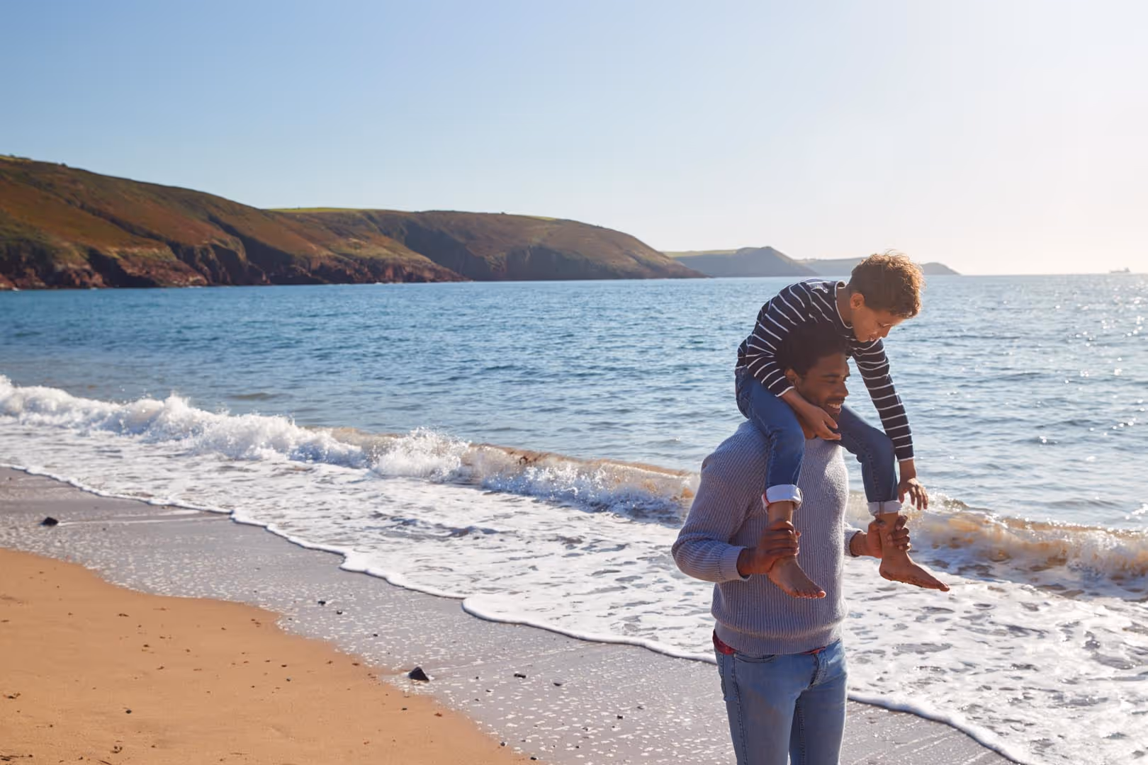 A man carries a child on his shoulders while walking along the beach near the ocean.