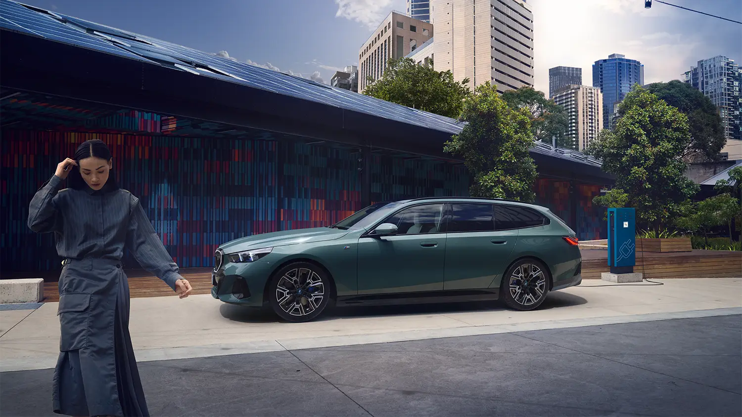 Ein eleganter, grün lackierter BMW Touring wird an einer Ladesäule geladen, während eine stilvoll gekleidete Frau vorbeigeht – MOTORHEIMAT Wagner-Schwarz in Kirchseeon, Traunstein, Altenmarkt, Wasserburg und Oberhaching