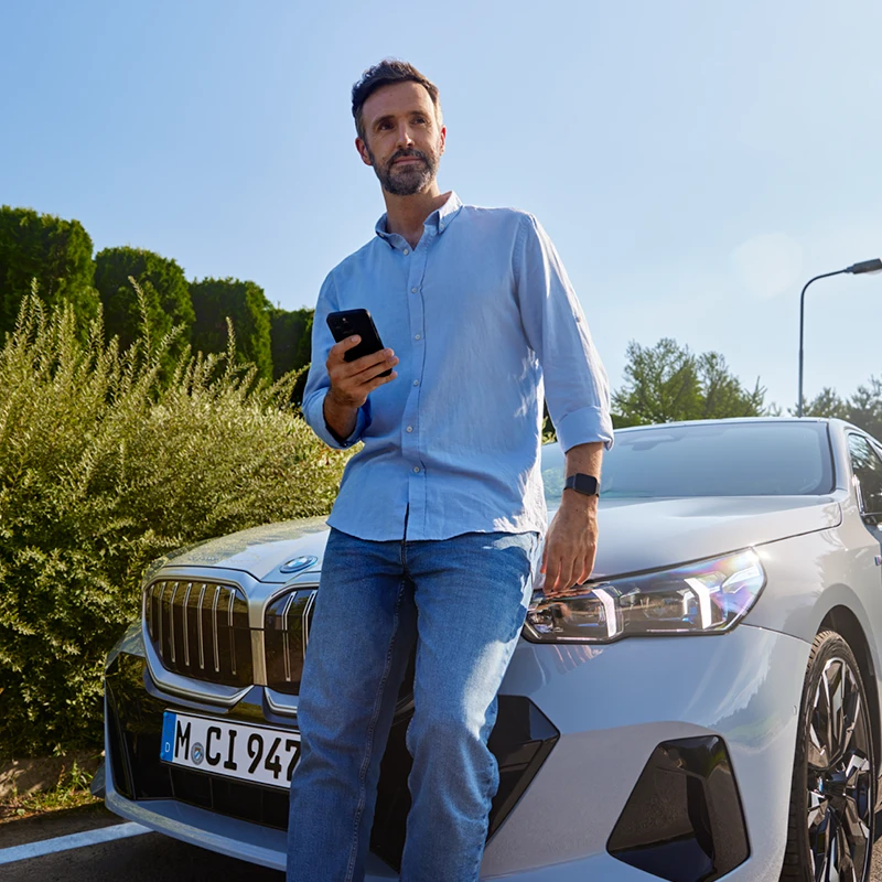 Mann in Jeans vor BMW i5 mit Smartphone in der Hand. Proactive Care in der MOTORHEIMAT Wagner-Schwarz