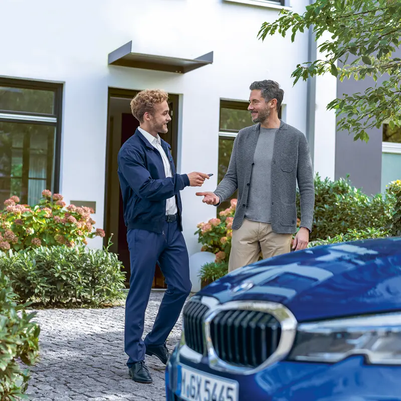 Zwei Männer vor einem Haus mit einem blauen BMW, welche sich einen Schlüssel übergeben.
