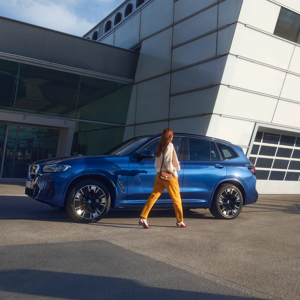 Blauer BMW iX3 vor einer Werkstatt mit einer Frau davor.