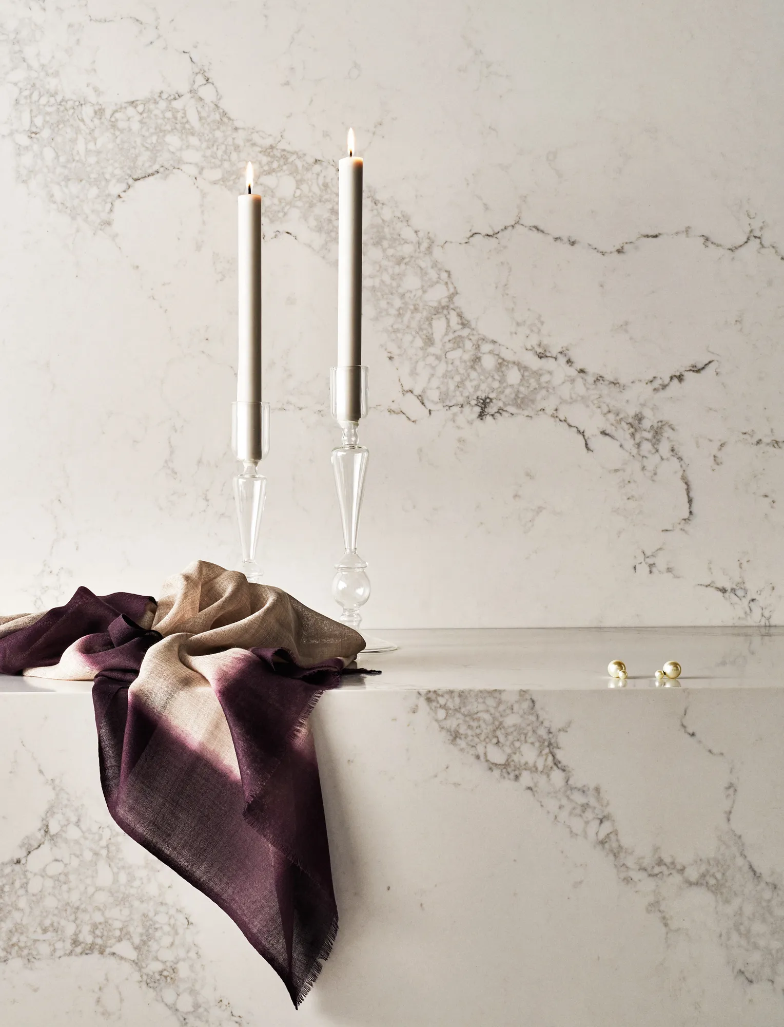 Two white lit taper candles in glass holders on a marble surface with a purple and beige cloth and pearl earrings nearby.