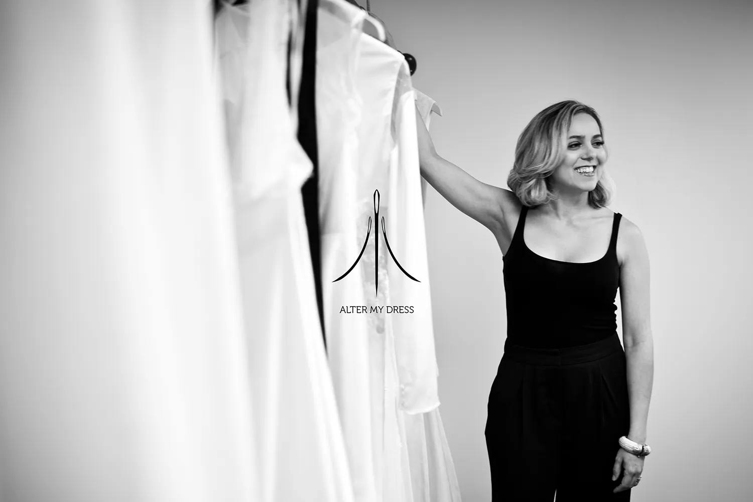 Alter My Dress logo on black and white background image of lead designer Priscilla smiling with wedding dresses on a rack