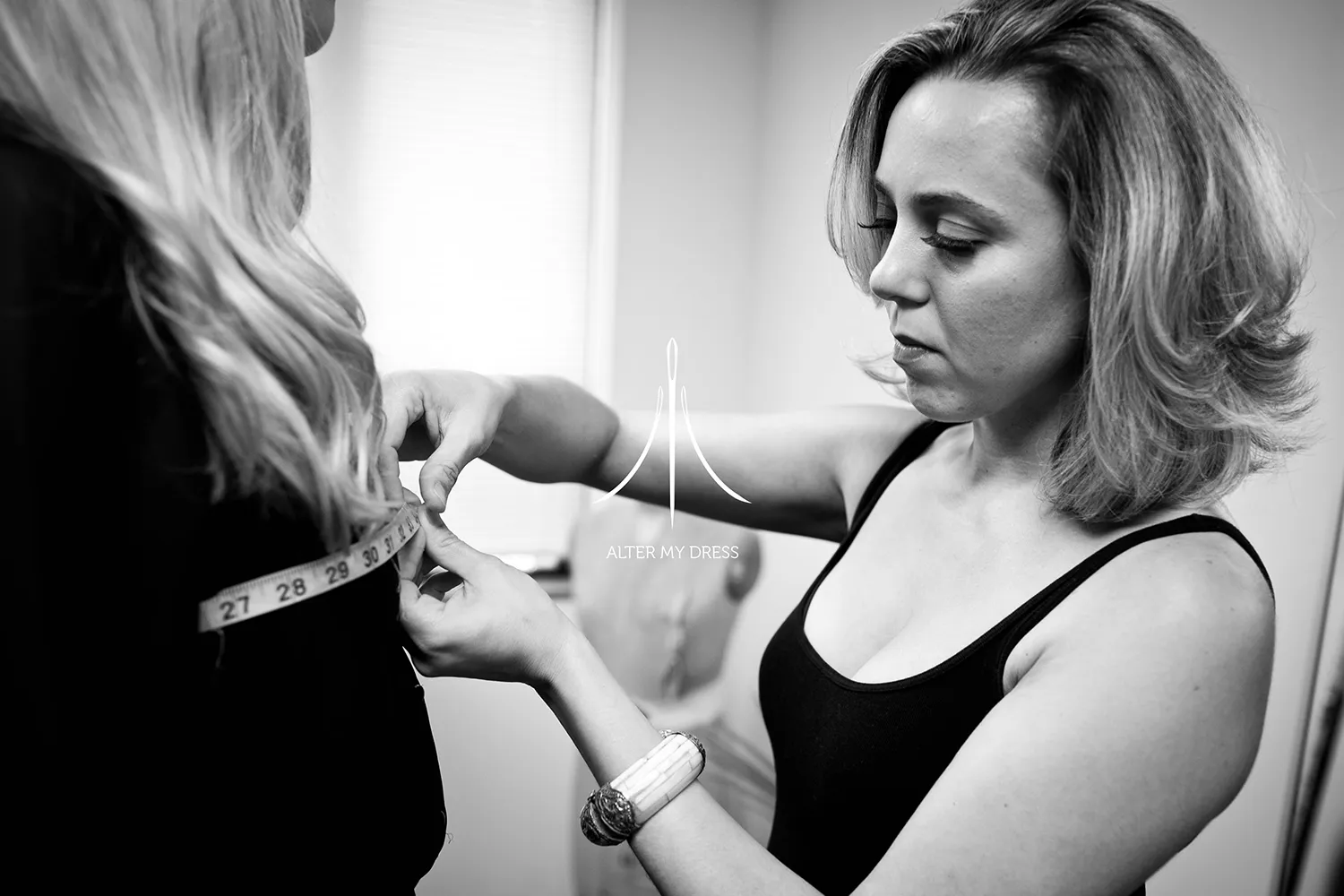 Fashion designer adjusting a sheer, beaded top and white tulle skirt worn by a model against a white background with Alter My Dress logo.