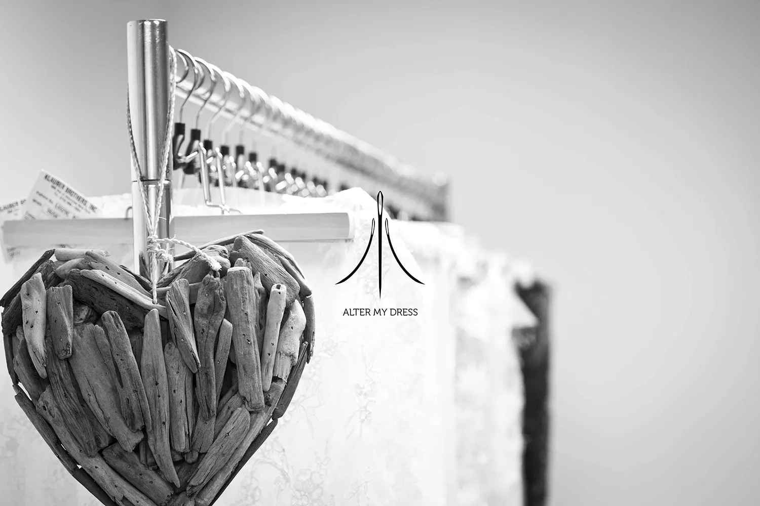 Alter My Dress logo on black and white background image of wedding dress alterations on a rack with a hanging heart made out of wood