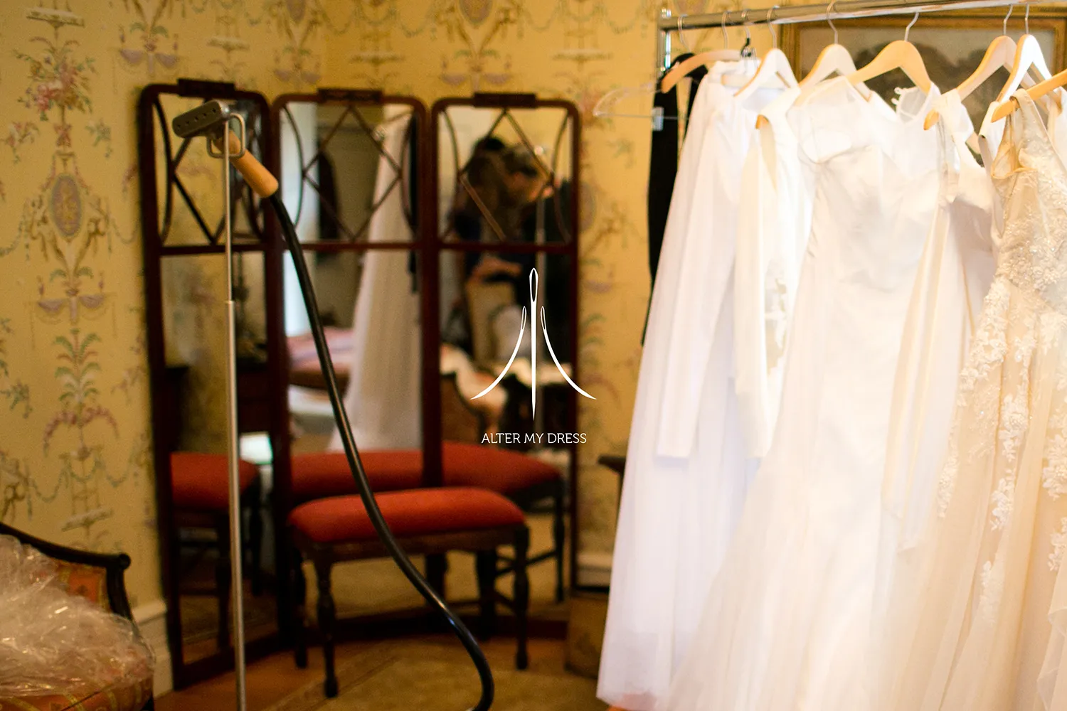 Steamer and a rack of white wedding dresses in a vintage-style room with mirrors and floral wallpaper.