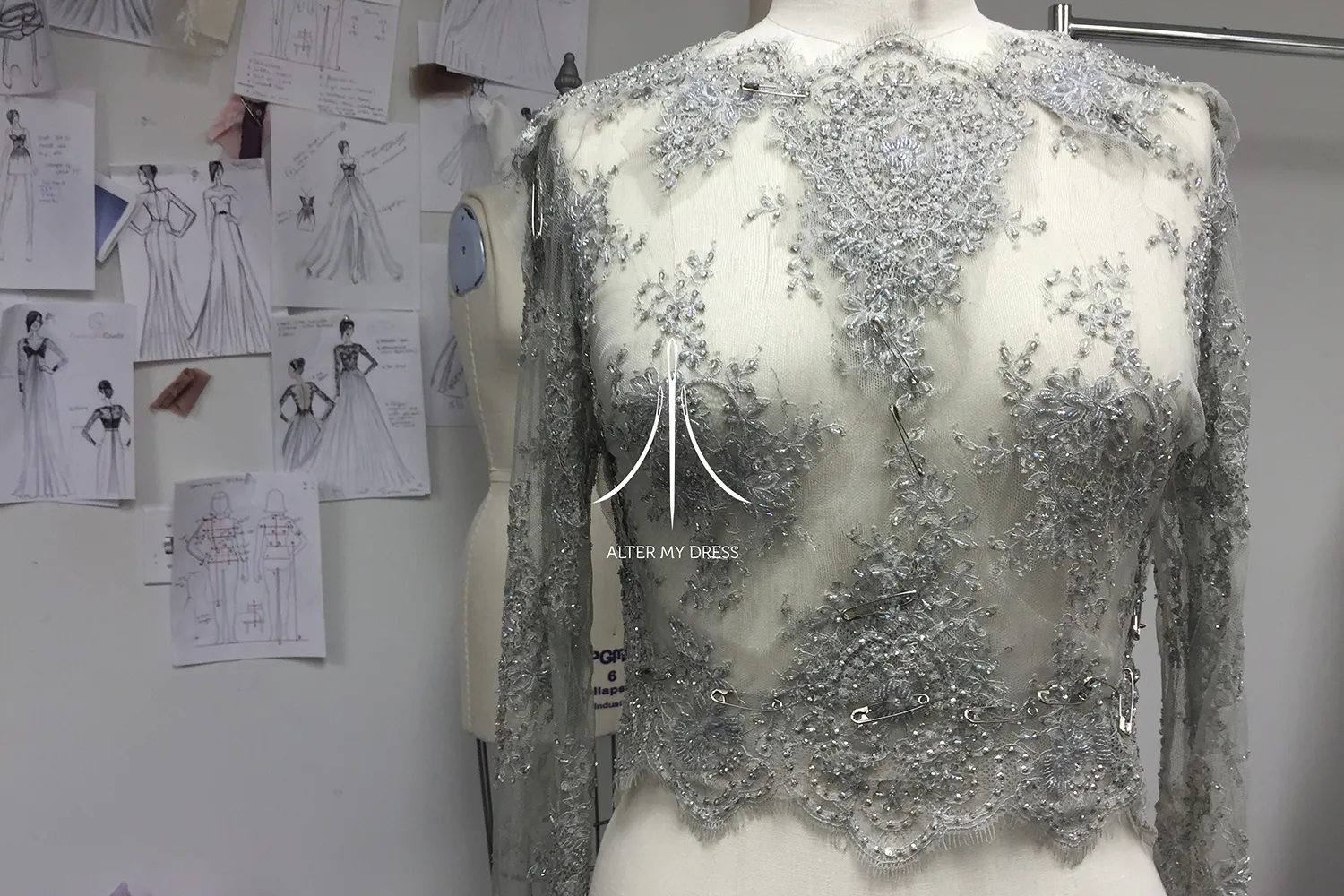 Close-up of a mannequin wearing a sheer, embellished lace bodice pinned for fitting, with fashion sketches on the wall behind.