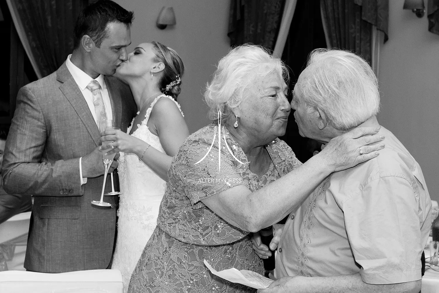 Black and white photo of a bride and groom kissing beside an elderly couple greeting each other with a kiss on the cheek.