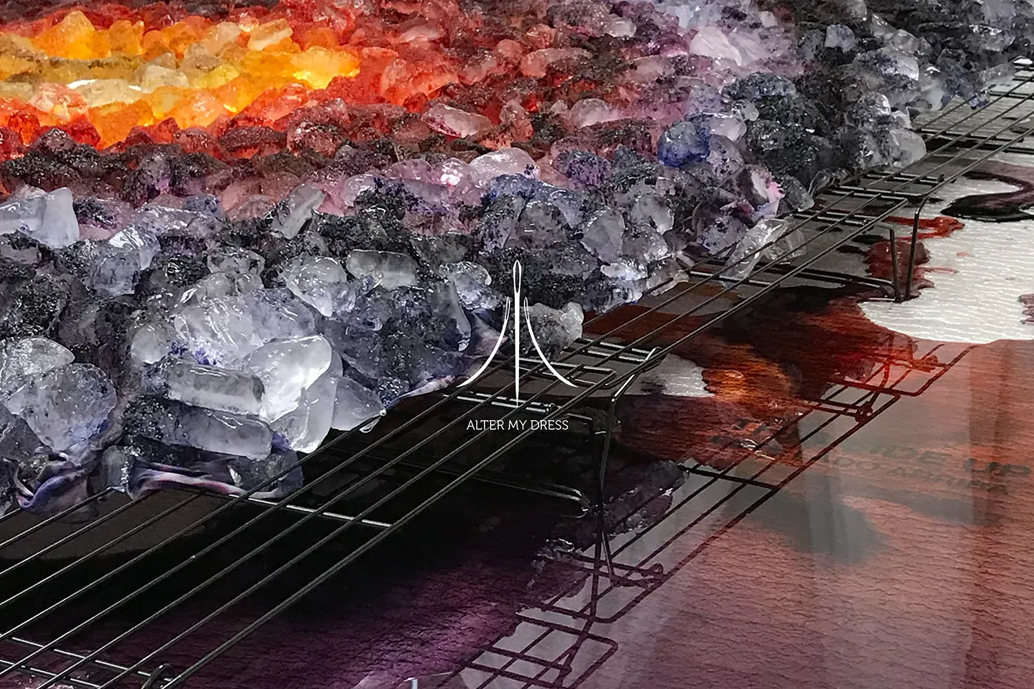 Close-up of colorful crushed ice in shades of orange, yellow, purple, and black on a metal rack with 'Alter My Dress' logo overlay.