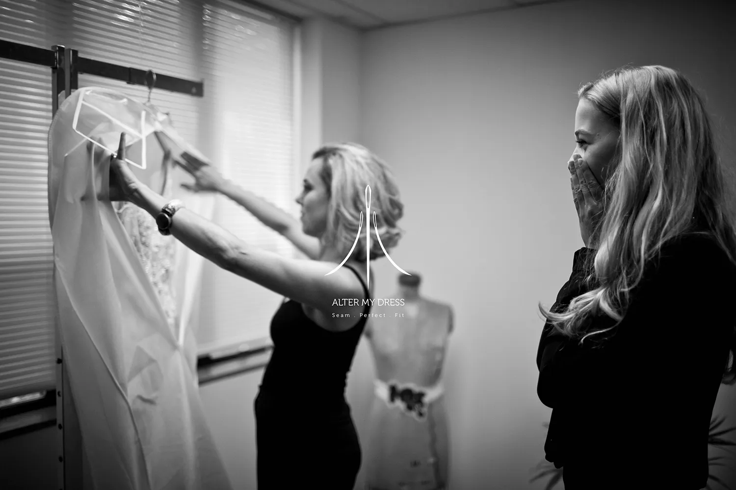 Two women examining a wedding dress in a fitting room, one holding the dress on a hanger and the other looking excited with hands covering her mouth.