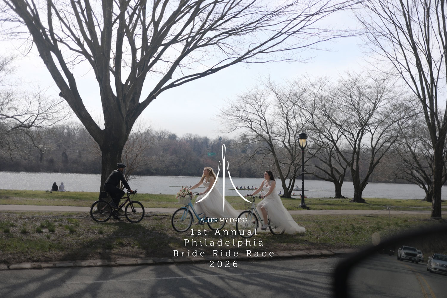 Two brides in wedding dresses riding bikes by a riverside park with leafless trees, accompanied by a cyclist.