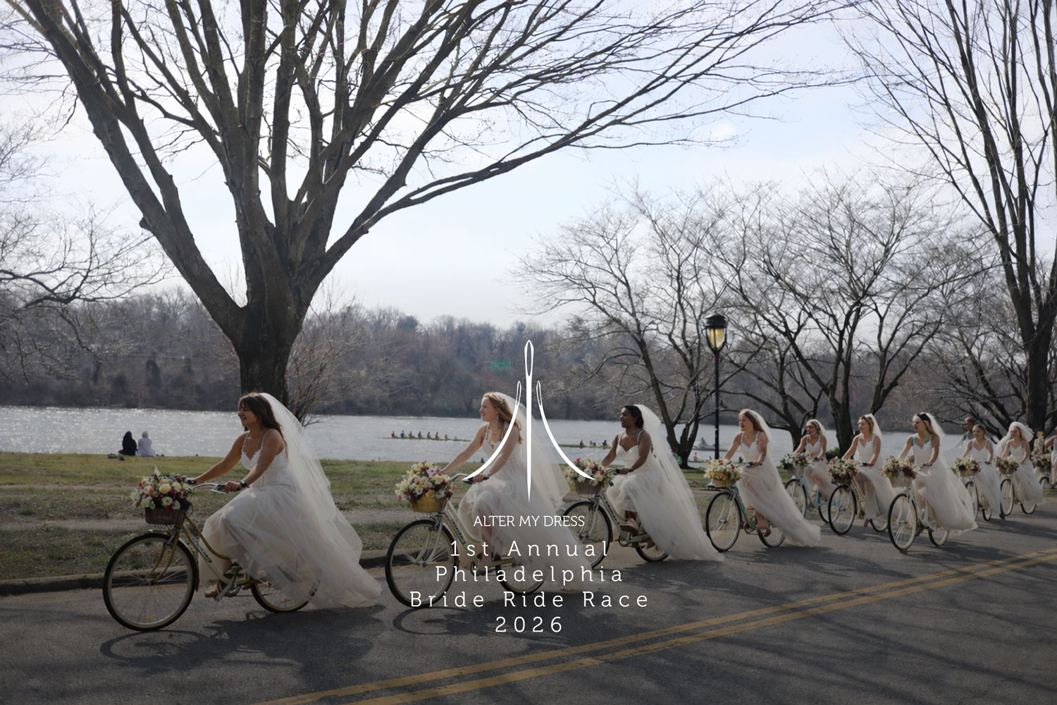 A line of brides in wedding dresses riding decorated bicycles along a tree-lined road by a river.