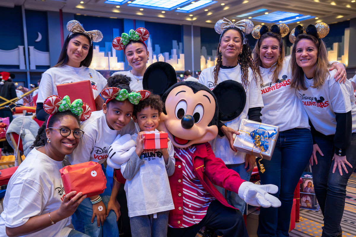 Des volontaires Disney portant des t-shirts "Disney VoluntEARS" posent avec Mickey et des enfants, tenant des cadeaux emballés lors d’un événement solidaire de Noël.