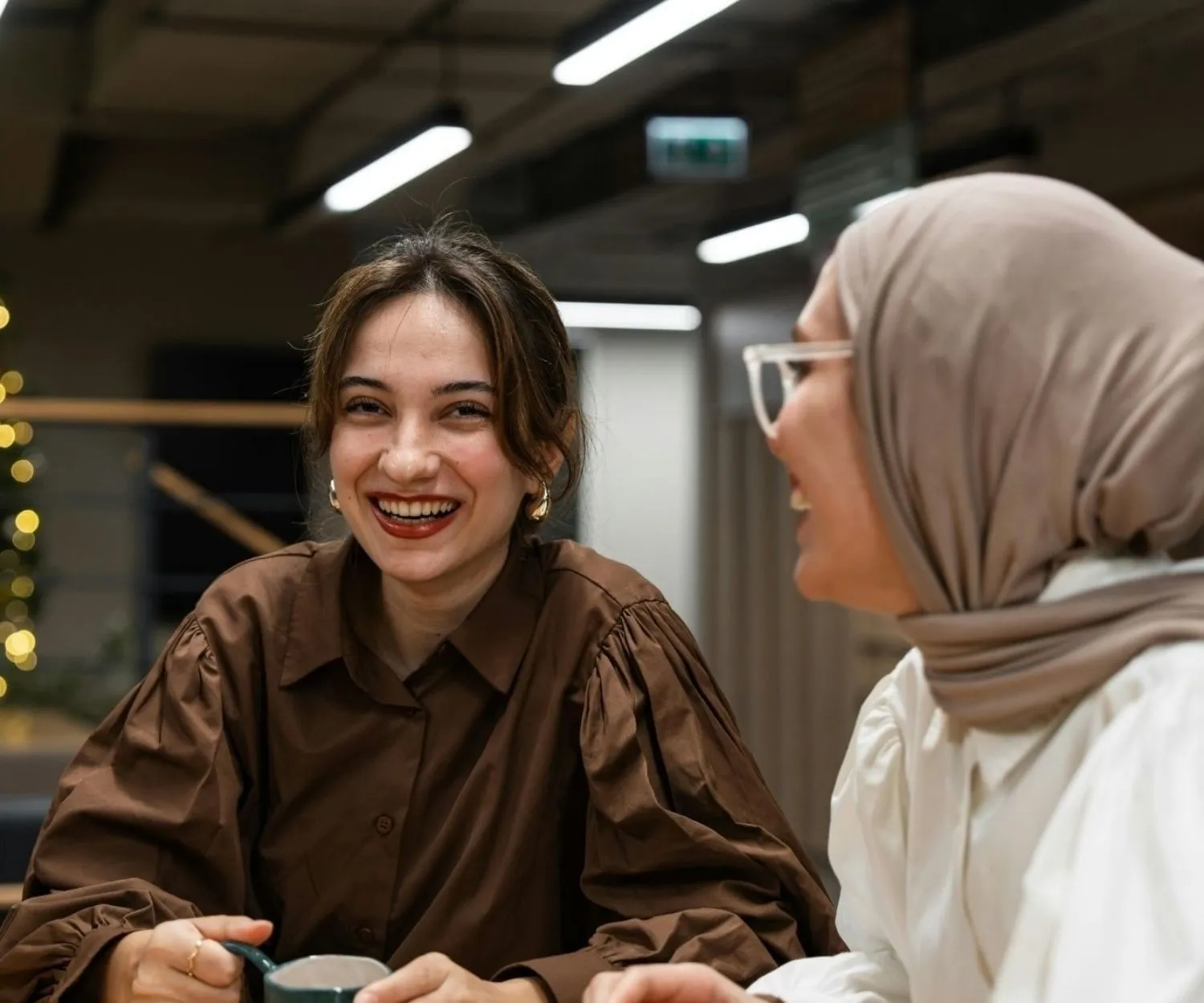 Deux femmes échangent autour d’une table dans un espace de travail moderne, créant un moment convivial et complice en intérieur.