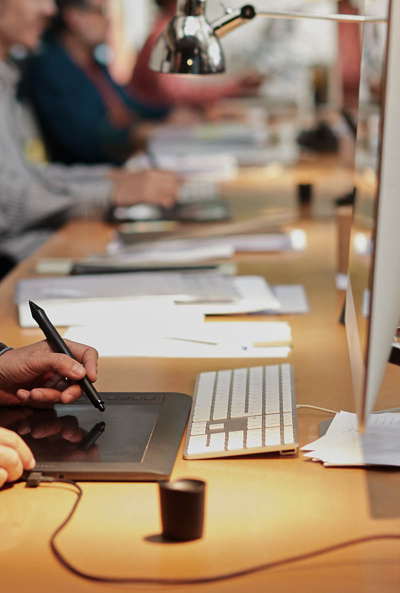 A hand holding a stylus over a graphic tablet on a desk with a keyboard and papers.