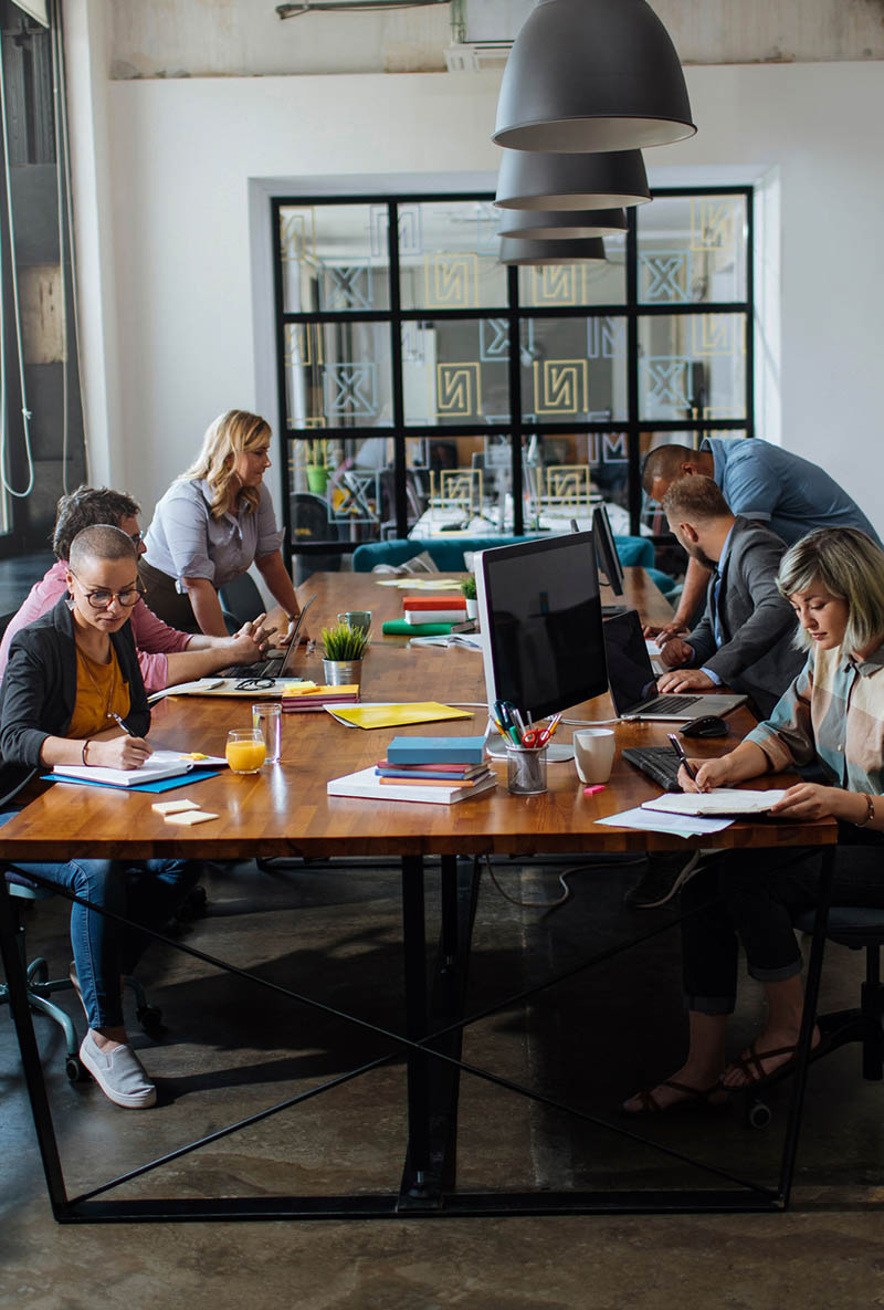 A team collaborating at a large wooden table, with laptops, notebooks, and drinks.