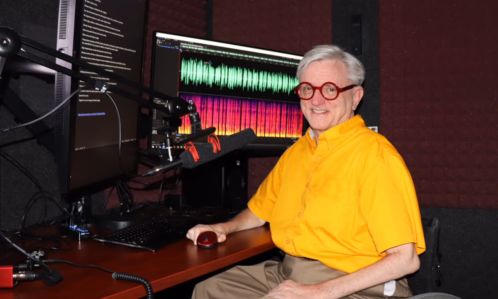 A narrator seated next to his recording station inside of a prefab vocal booth by WhisperRoom.