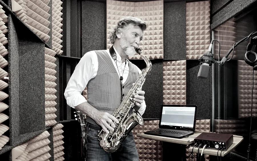 A saxophonist plays into a mounted microphone inside a foam-lined WhisperRoom, with a laptop on a stand beside him