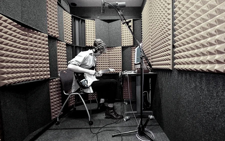 A guitarist on a stool plays an electric guitar into a microphone inside a foam-lined WhisperRoom booth