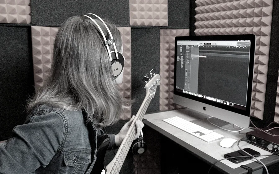 A bass player wearing headphones tracks a part inside a WhisperRoom, watching the recording session on a desktop screen