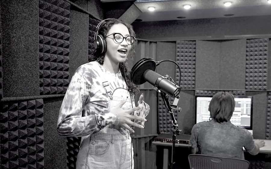 A singer wearing headphones performs into a studio microphone inside a WhisperRoom lined with acoustic foam, with an engineer at a screen behind her