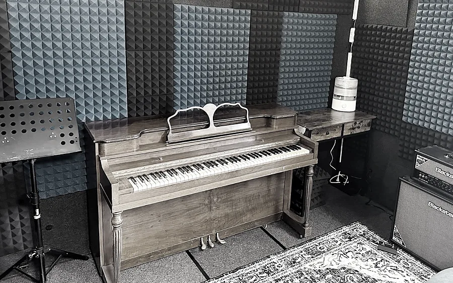 An upright piano and a music stand inside a WhisperRoom with foam-treated walls and a rug on the floor