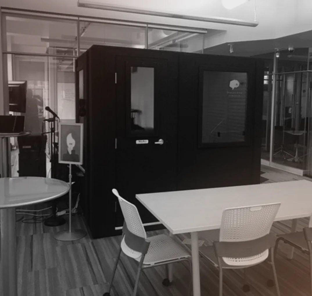 A customer's WhisperRoom booth installed in an open-plan office, next to a meeting table and chairs, with glass-walled offices behind it