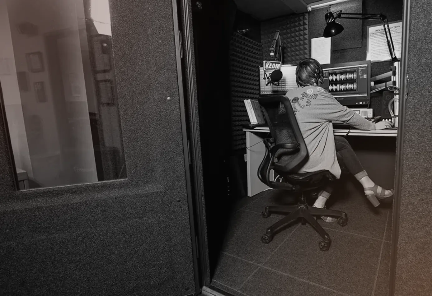 A KEOM radio host on air at the station's console inside a WhisperRoom broadcast booth, seen past the booth's open door and its window