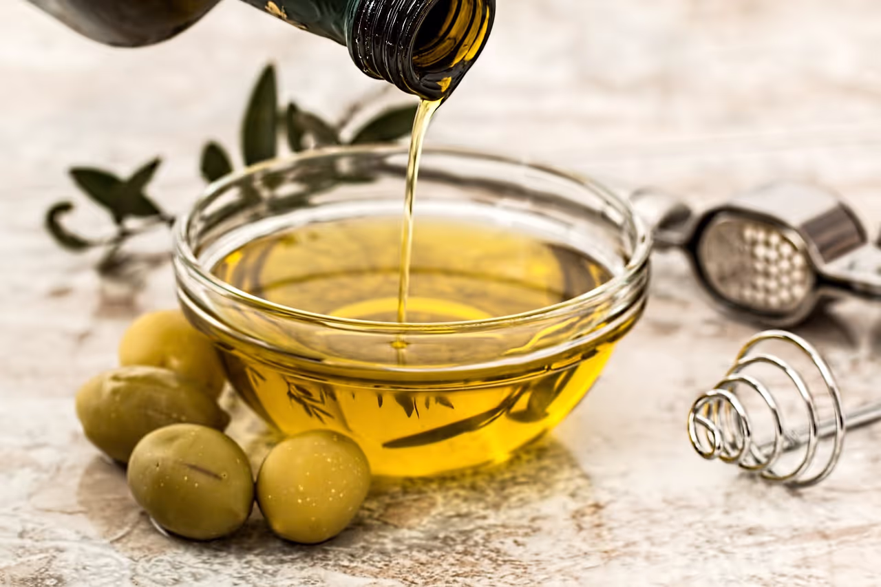 Olive oil being poured into a glass bowl surrounded by green olives and a metal kitchen utensil on a marble surface.