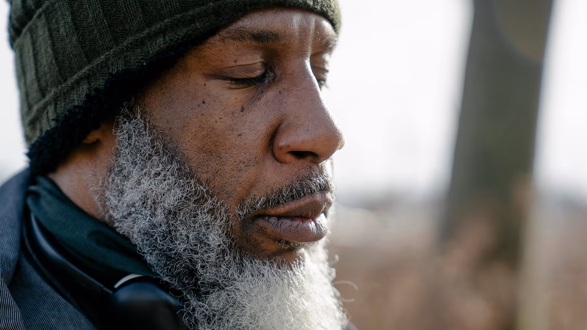 Close-up of a contemplative older man with a gray beard wearing a green knit hat and a dark jacket outdoors.