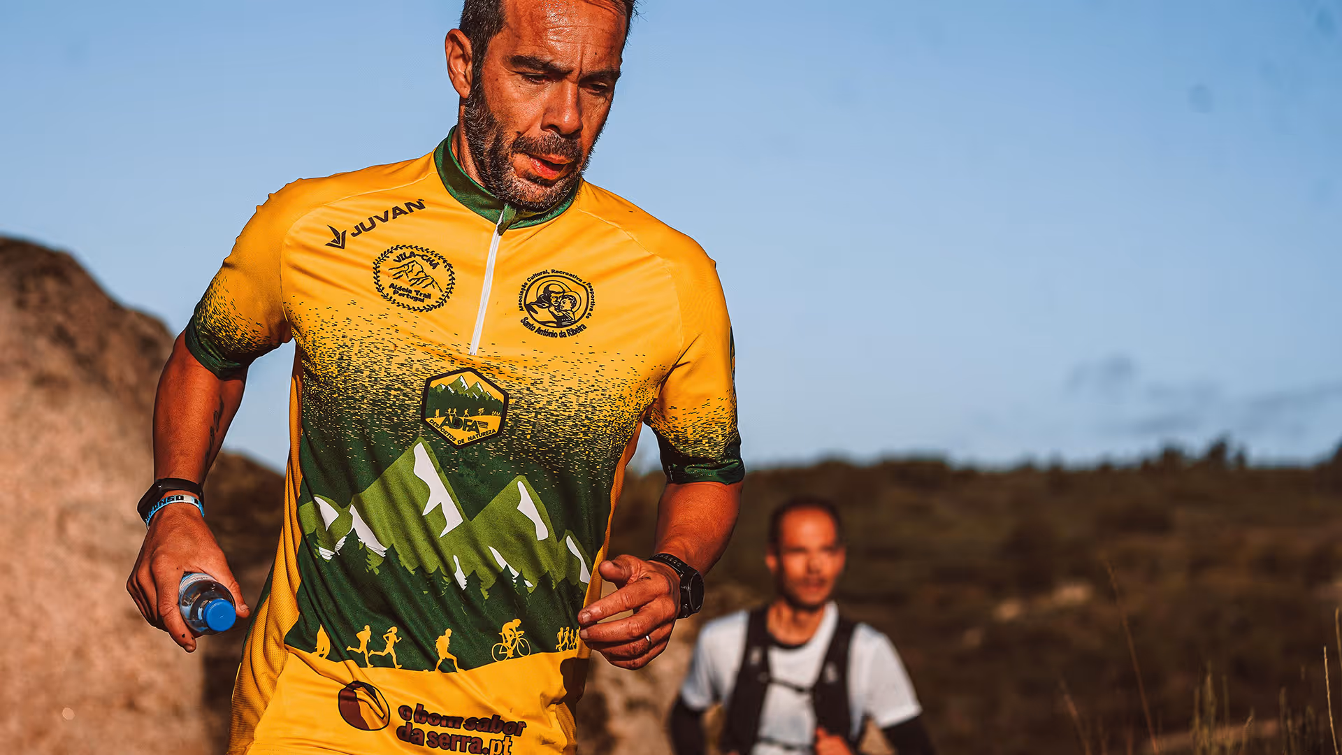 Man in yellow and green athletic shirt running outdoors holding a water bottle, with another runner in background.
