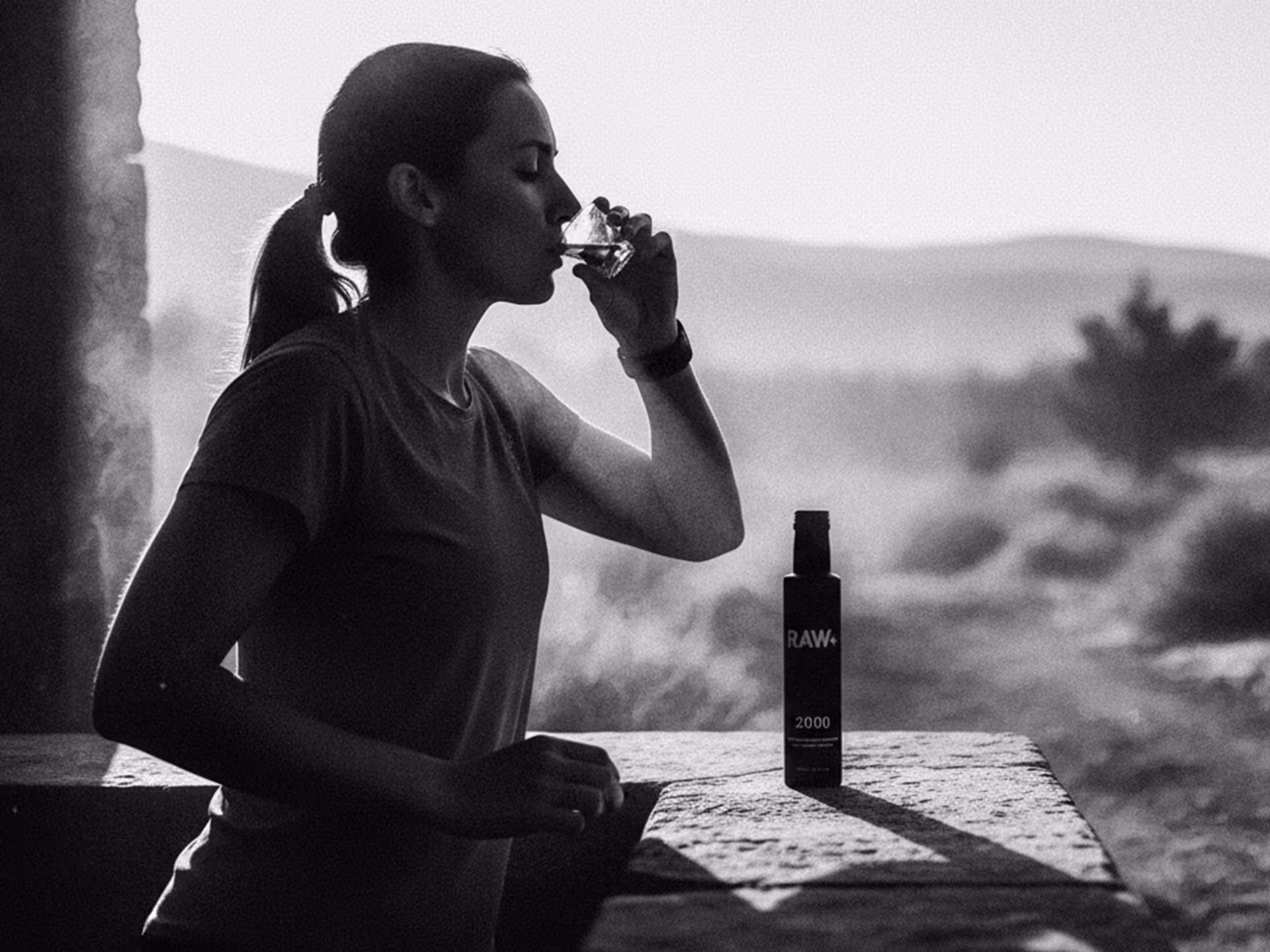 Woman drinking from a small glass while standing by a stone ledge with a RAW 2000 bottle on it, outdoors with blurred natural background.