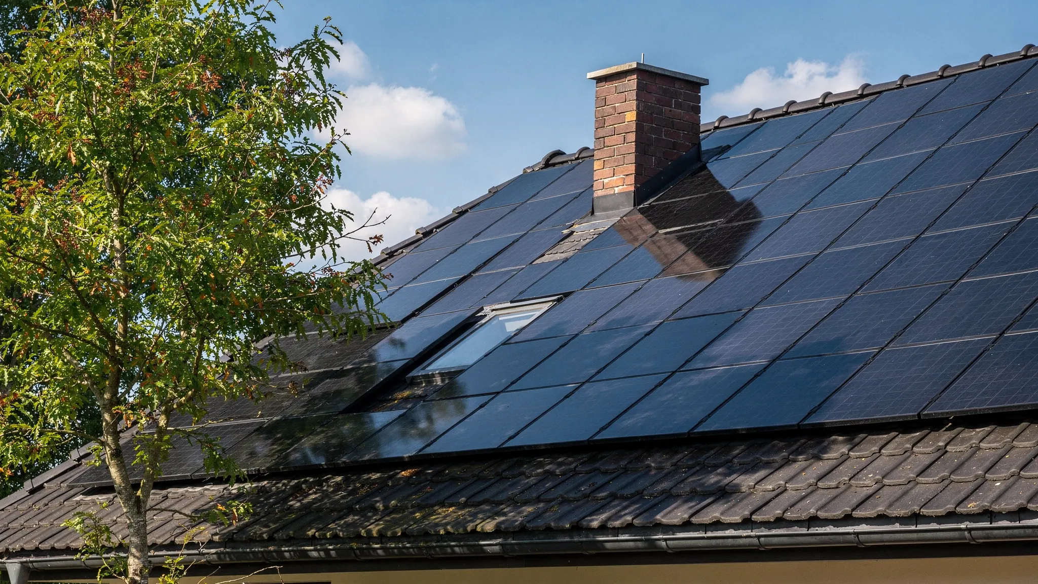 Einfamilienhausdach mit Photovoltaikmodulen, sichtbarem Schornstein und einem nahe stehenden Baum, der am Rand des Dachs Schatten wirft. Die Szene verdeutlicht, wie Verschattung und Dachdetails Ertragsprognosen beeinflussen.