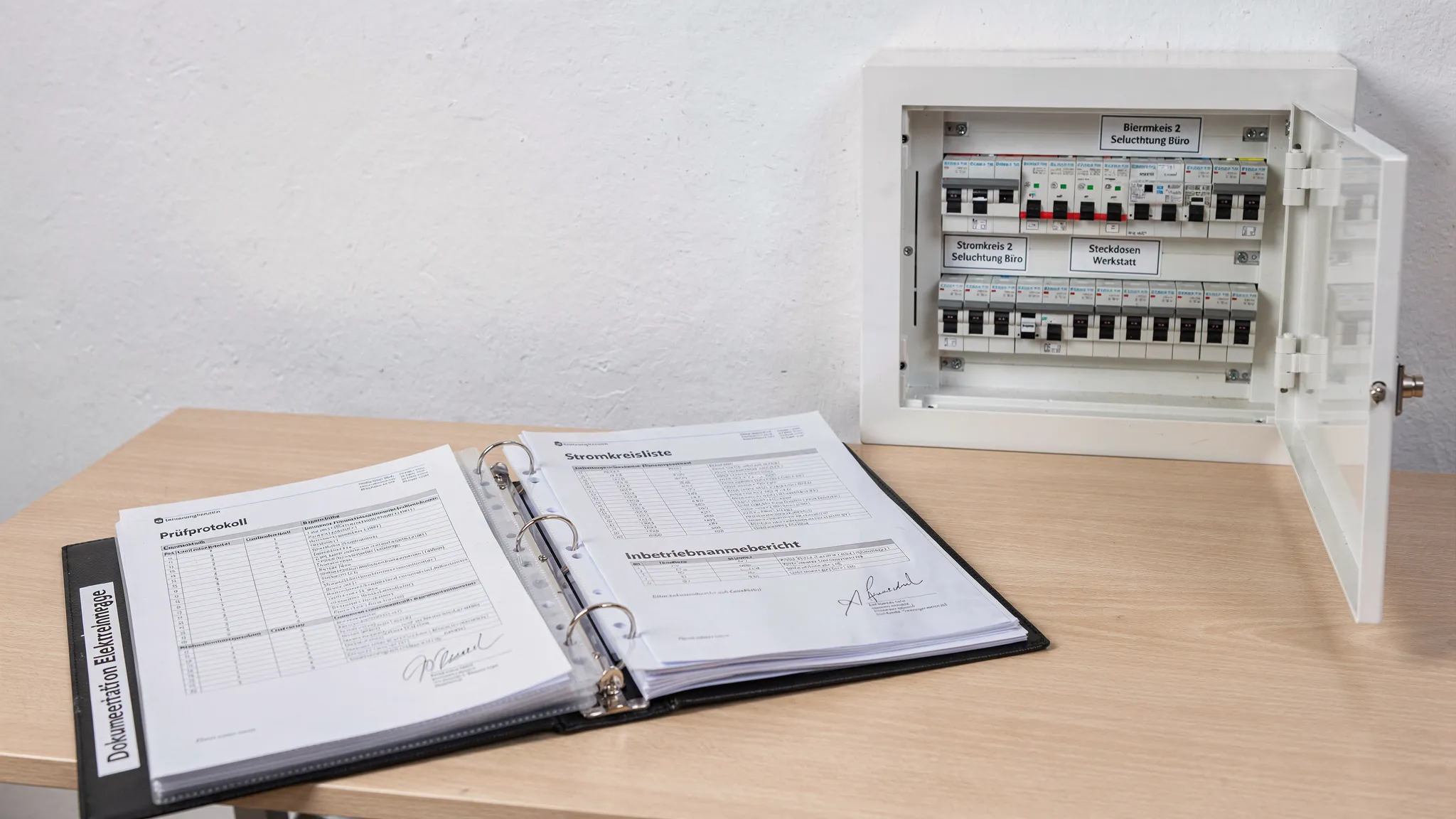 Ein aufgeräumter Dokumentationsordner mit Prüfprotokollen, Stromkreislisten und Inbetriebnahmeberichten liegt neben einem beschrifteten Sicherungskasten; alle Unterlagen sind sauber abgeheftet.