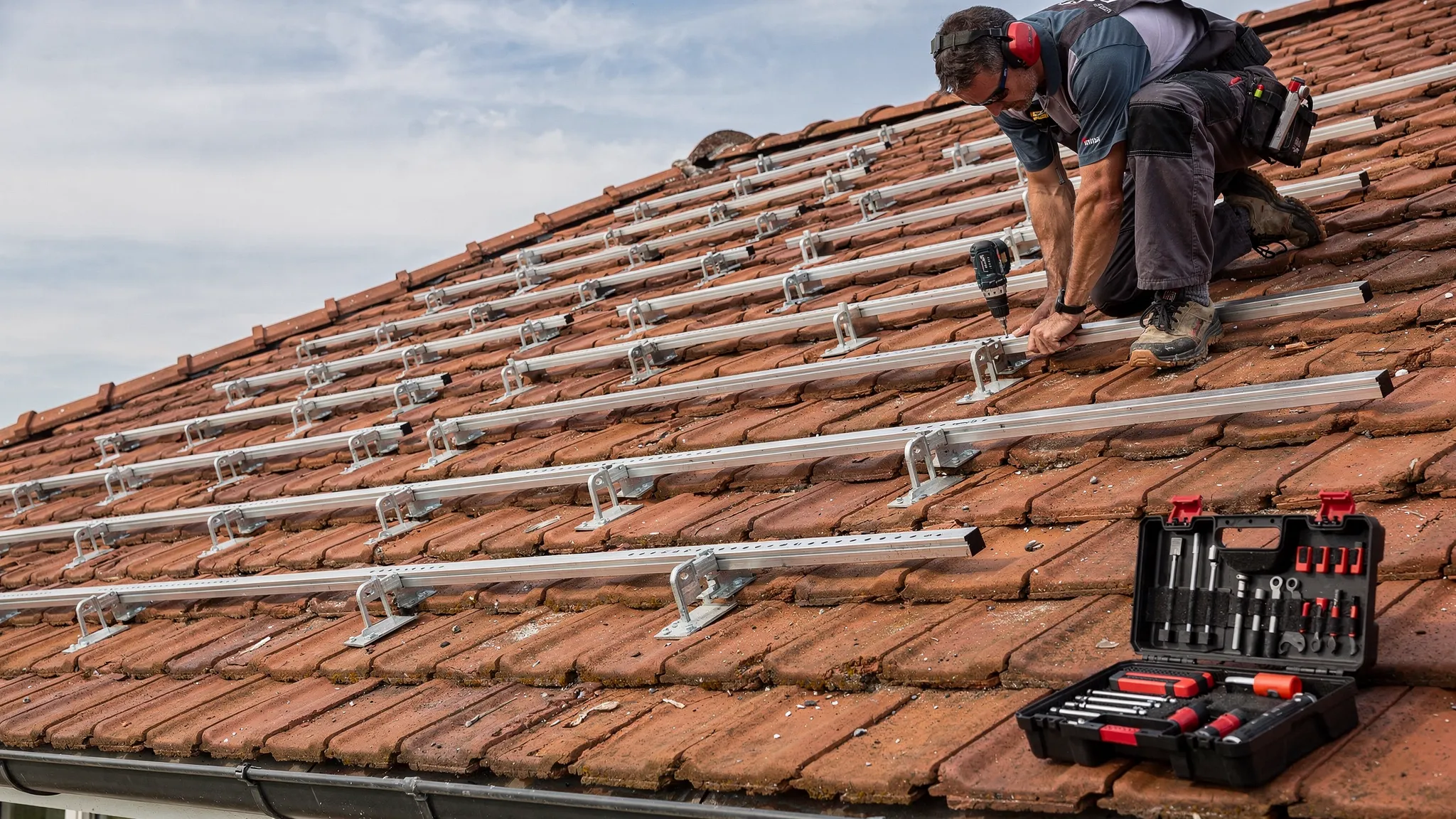 Ein Installateur befestigt ein Montagesystem auf einem geneigten Ziegeldach; zu sehen sind Dachhaken, Montageschienen und ein Werkzeugkoffer am Dachrand.