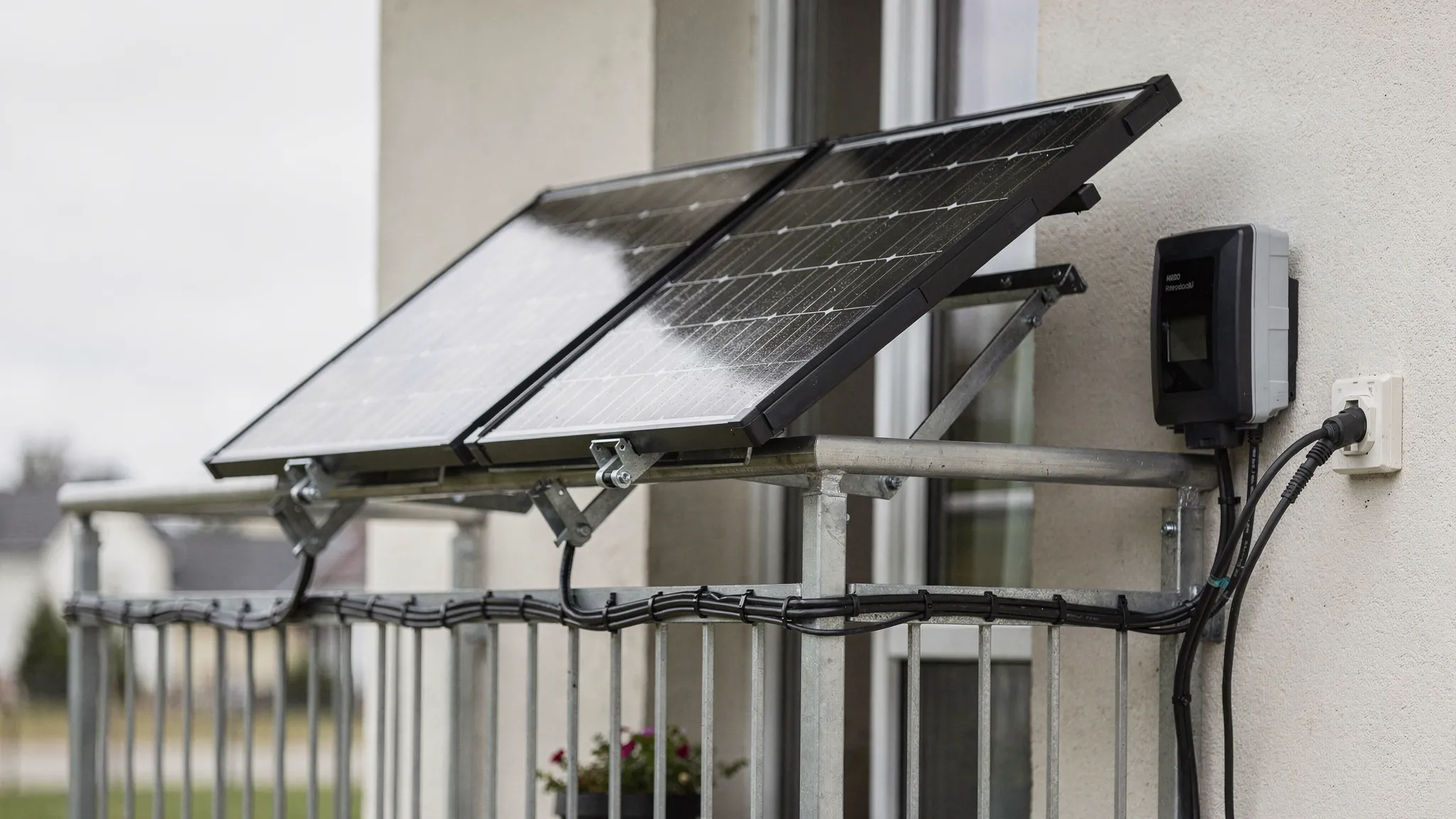 Zwei Solarmodule sind sicher an einem Balkon montiert, daneben sieht man wetterfeste Kabel, einen kleinen Wechselrichter und eine Außensteckdose an der Hauswand.