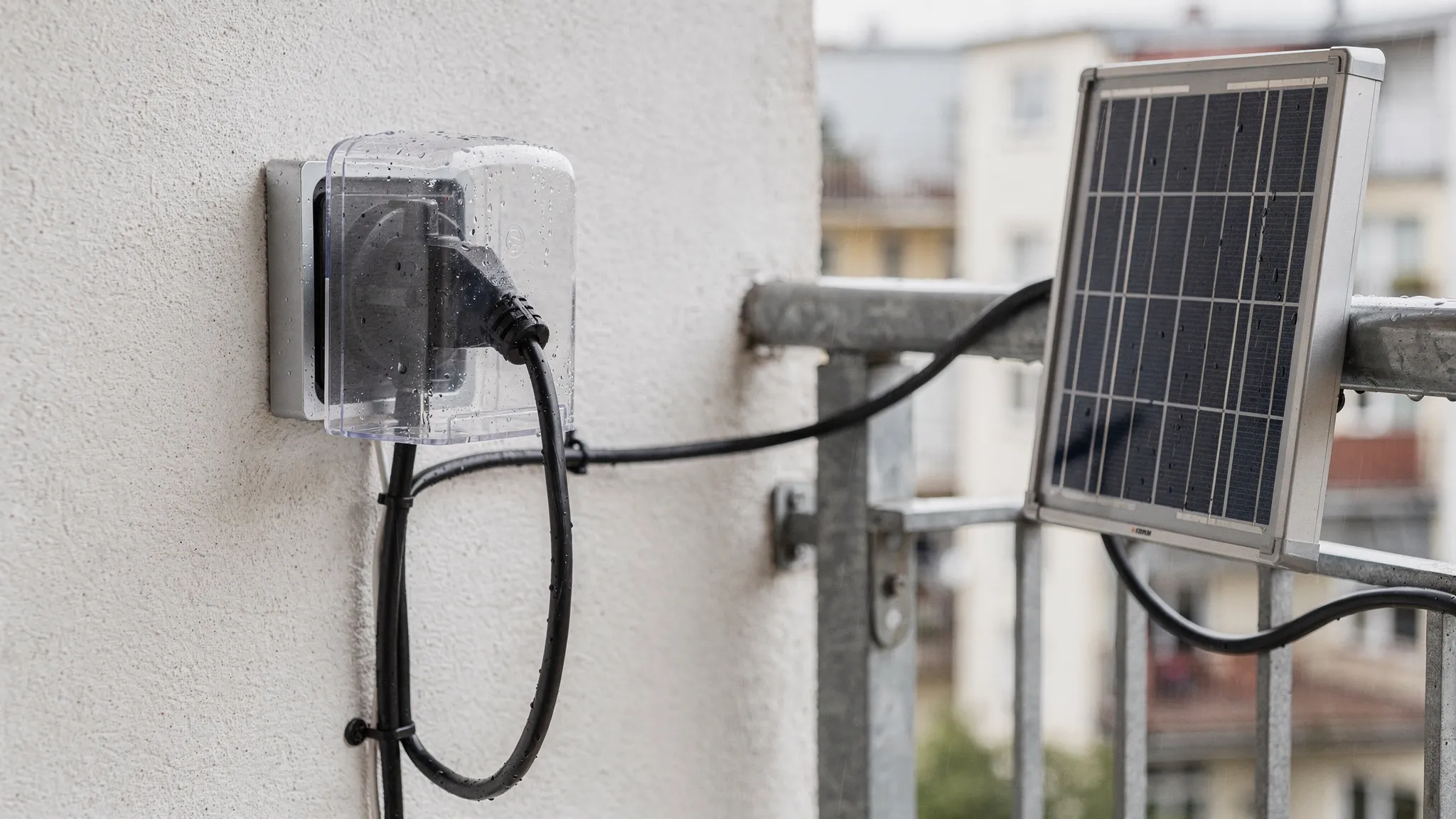 Eine wettergeschützte Außensteckdose an einer Balkonwand mit eingestecktem Kabel einer Balkon-Solaranlage, sauber geführte Solarkabel mit Tropfschleife und ein kleines Solarmodul am Balkongeländer.