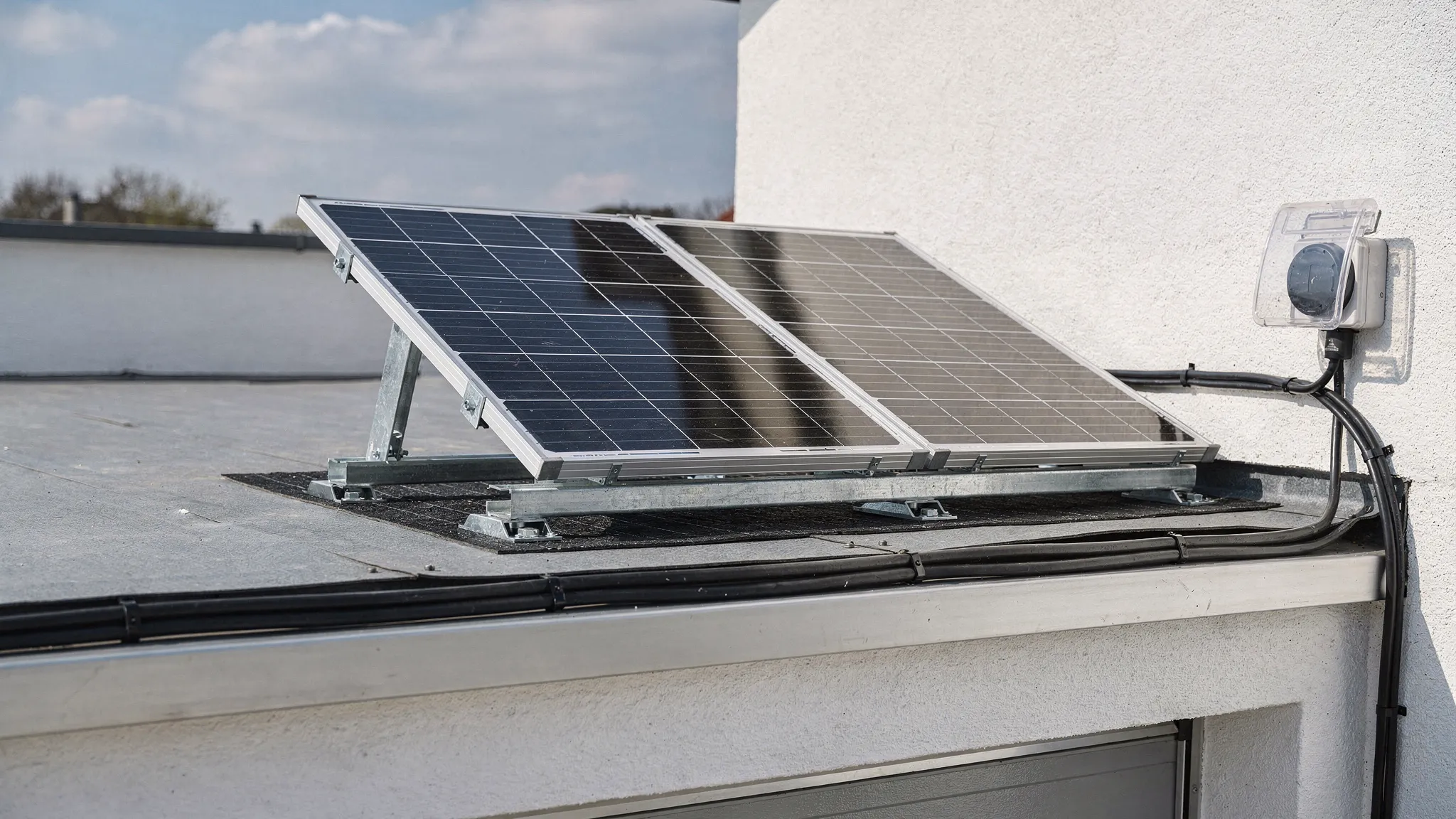 Ein kleines Balkonkraftwerk mit zwei Solarmodulen auf einem flachen Garagendach, montiert auf ballastierten Flachdachständern mit Schutzmatten unter der Unterkonstruktion. Die Module stehen mit Abstand zum Dachrand, Kabel sind ordentlich befestigt und führen zu einer wettergeschützten Außensteckdose.