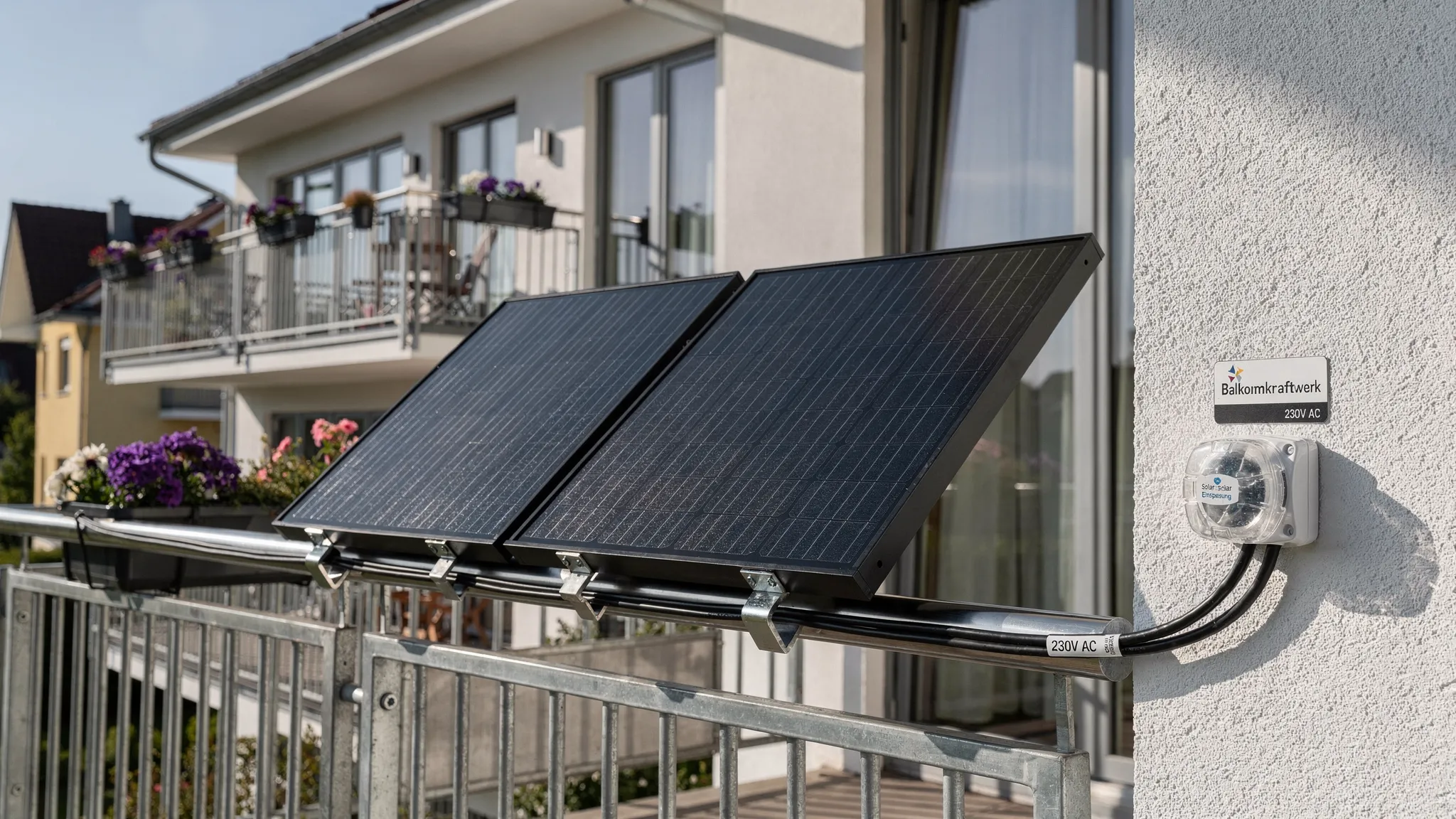 Zwei kompakte Solarmodule sind sicher an einem sonnigen Balkon montiert. Im Hintergrund sieht man ein Wohnhaus mit Terrasse und unverschatteter Südausrichtung, während die Kabel sauber und geschützt zur Außensteckdose geführt werden.