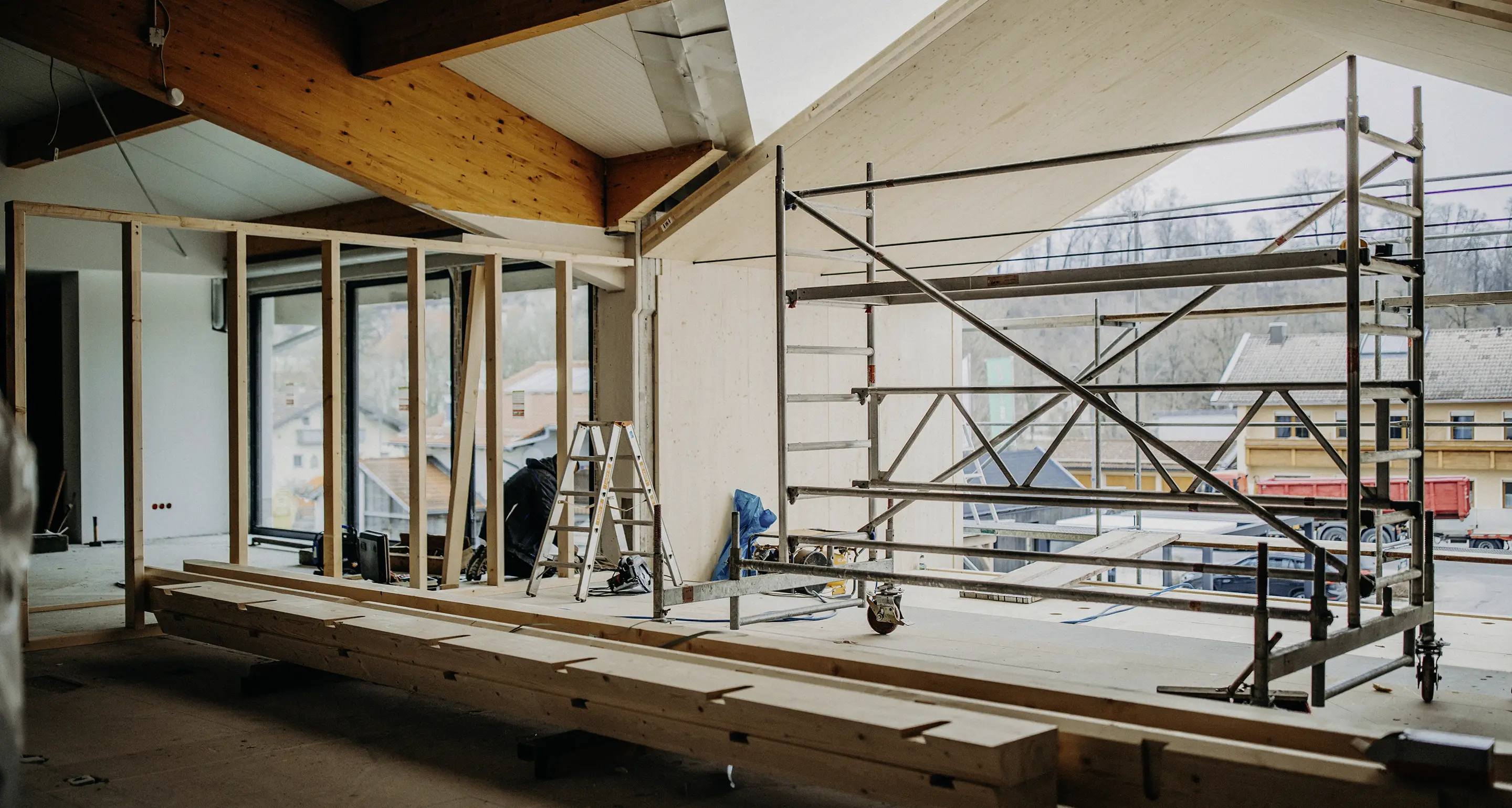 Innenansicht einer Baustelle mit Holzbalken, Gerüst und Leiter, mit Blick auf benachbarte Gebäude durch große Fenster.