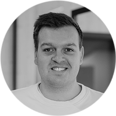 Kilian Heidl trägt weißen Pullover mit Aufdruck und Jeans. Er steht vor Glastür und trägt Armbanduhr am linken Handgelenk.