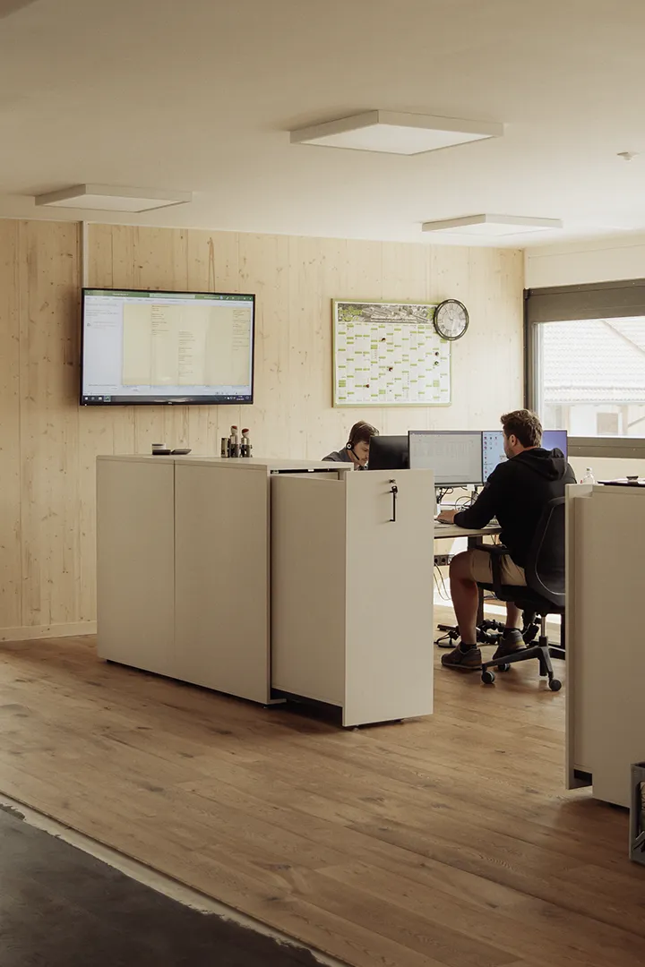 Zwei Personen arbeiten in einem modernen Büro mit Holzverkleidung an ihren Computerarbeitsplätzen; auf einem großen Bildschirm an der Wand sind Pläne und Dokumente zu sehen.