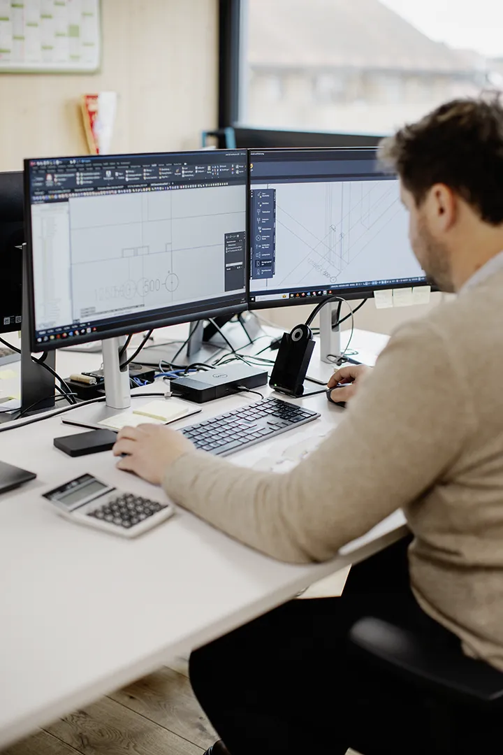 Ein Ingenieur arbeitet am Computer an technischen Zeichnungen und 3D-Modellen auf zwei Monitoren; auf dem Schreibtisch liegen Taschenrechner und Notizen.