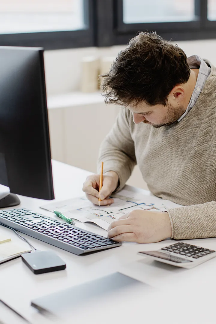 Ein Ingenieur arbeitet am Computer an technischen Zeichnungen und 3D-Modellen; auf dem Schreibtisch liegen Taschenrechner und Notizen.