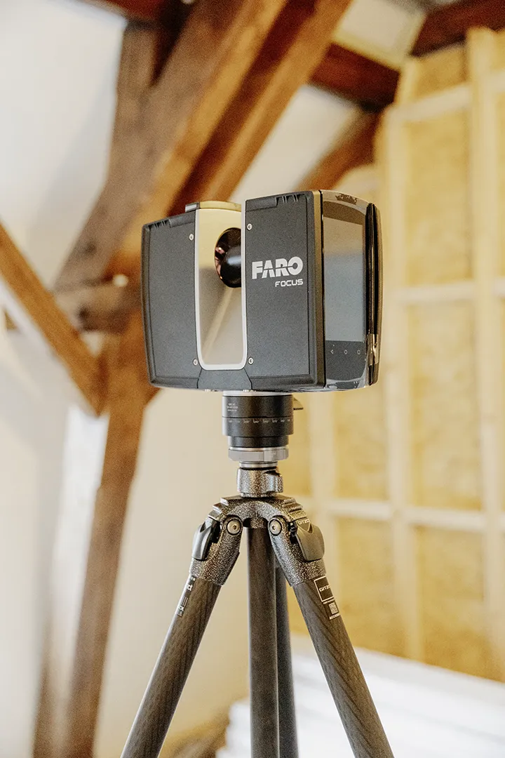 Ein FARO-3D-Laserscanner steht auf einem Stativ in einem Innenraum mit sichtbaren Holzbalken und Dämmung, um präzise Aufmaße im Bereich Innenausbau oder Trockenbau zu erfassen.